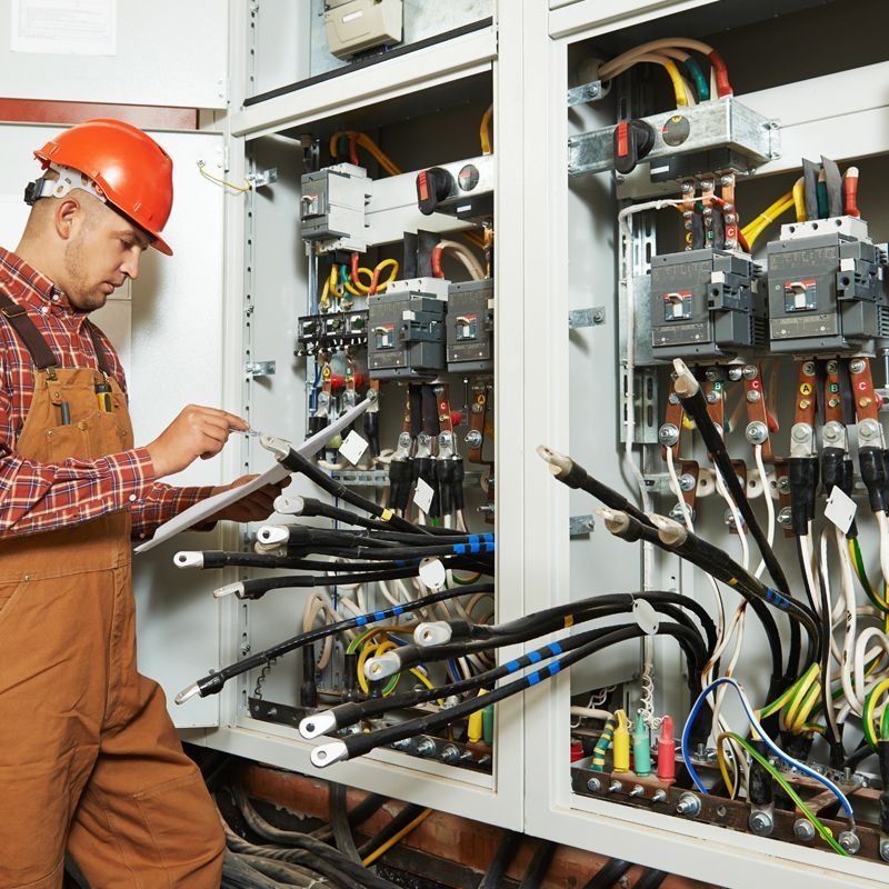 Arbeiter in orangefarbener Schutzausrüstung steht vor einem großen elektrischen Schaltkasten und überprüft die Ausrüstung